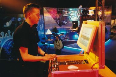Das Bild zeigt einen jungen Mann an einem Steuerpult mit Monitor und Bedienfeld, vermutlich zur Steuerung von Licht oder Audio, auf einer Messe oder Veranstaltung. Der bunte, beleuchtete Hintergrund und die Gestaltung vermitteln einen Retro-Charme aus den 1990er oder frühen 2000er Jahren.