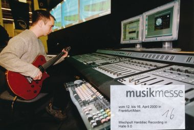 Das Bild zeigt einen Mann, der eine rote E-Gitarre spielt, während er vor einem großen Mischpult sitzt. Im Hintergrund sind zwei Monitore mit Audioprogrammen zu sehen, die den Aufnahmeprozess anzeigen. Die Szene hat auf einer Messe stattgefunden zu haben, genauer auf der Musikmesse vom 12. bis 16. April 2000 in Frankfurt am Main, wie die eingeblendete Beschriftung angibt. Das Setting zeigt eine Präsentation von Mischpulten und Harddisc-Recording-Technologie in Halle 9.