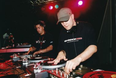 Das Bild zeigt zwei DJs, die an ihren Turntables arbeiten, während sie konzentriert in ihre Performance vertieft sind. Die Szene ist von stimmungsvollem Bühnenlicht in Rot- und Violetttönen beleuchtet, was die dynamische Atmosphäre unterstreicht. Beide DJs tragen Kopfhörer und schwarze T-Shirts mit Schriftzügen, die auf ihre Beteiligung an einem Event wie „Scratchathon 2000“ hinweisen. Im Vordergrund sind mehrere Plattenspieler und Mischpulte zu sehen, die eine professionelle DJ-Ausrüstung darstellen. Im Hintergrund ist schemenhaft ein weiterer Performer zu erkennen. Das Bild vermittelt die Energie und Leidenschaft eines Live-DJ-Auftritts.
