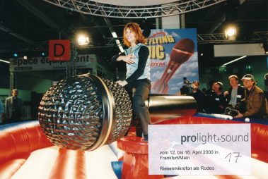 Das Bild zeigt eine Frau, die lachend auf einem mechanischen Rodeogerät in Form eines riesigen Mikrofons sitzt. Die Szene spielt auf der Prolight + Sound Messe, die vom 12. bis 16. April 2000 in Frankfurt am Main stattfand, wie der Text auf dem Bild verrät. Im Hintergrund sind ein Banner mit der Aufschrift „The Flying Mic“ und weitere Besucher der Messe zu sehen. Das riesige Mikrofon ist detailliert gestaltet und in eine rot-blau-weiße aufblasbare Fläche integriert, die als Sicherheitsbereich dient. Die Szenerie wird von Kameraleuten und Messebesuchern beobachtet, was die interaktive und unterhaltsame Atmosphäre des Events unterstreicht.