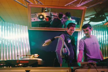 Das Bild zeigt zwei Männer, die sich vor einem Mischpult mit DJ-Equipment, einschließlich eines Vestax-Plattenspielers, unterhalten. Ein gerahmter Bildschirm oben spiegelt die Technik. Der Hintergrund ist bunt beleuchtet und industriell gestaltet, typisch für eine Messe der 1990er oder 2000er Jahre.