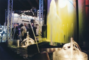 Das Bild zeigt ein beeindruckendes Farbenspiel aus sich kreuzenden Lichtstrahlen, die in verschiedenen leuchtenden Farben wie Grün, Blau, Gelb und Violett strahlen. Die Strahlen scheinen von einem zentralen Punkt auszugehen, wodurch ein symmetrisches Muster entsteht, das eine dynamische und fast hypnotische Wirkung erzeugt. Die dunkle Umgebung verstärkt die Intensität der leuchtenden Farben und lässt die Lichtstrahlen besonders hervorstechen. Diese Szene könnte Teil einer Lichtshow oder einer Lasertechnologie-Demonstration sein, die darauf abzielt, die ästhetische und technische Faszination moderner Beleuchtungssysteme zu präsentieren.