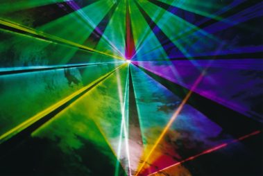 Das Bild zeigt ein beeindruckendes Farbenspiel aus sich kreuzenden Lichtstrahlen, die in verschiedenen leuchtenden Farben wie Grün, Blau, Gelb und Violett strahlen. Die Strahlen scheinen von einem zentralen Punkt auszugehen, wodurch ein symmetrisches Muster entsteht, das eine dynamische und fast hypnotische Wirkung erzeugt. Die dunkle Umgebung verstärkt die Intensität der leuchtenden Farben und lässt die Lichtstrahlen besonders hervorstechen. Diese Szene ist Teil einer Lichtshow oder einer Lasertechnologie-Demonstration, die darauf abzielt, die ästhetische und technische Faszination moderner Beleuchtungssysteme zu präsentieren.