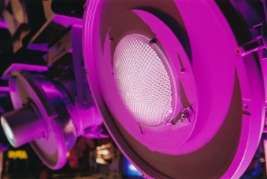 Das Bild zeigt ein Nahaufnahme einer professionellen Bühnenleuchte, die in ein intensives violettes Licht getaucht ist. Die Struktur der Linse ist gut erkennbar und unterstreicht die technische Präzision des Geräts. Im Hintergrund deuten unscharfe Elemente auf die Umgebung einer Messe oder Ausstellung hin, wahrscheinlich im Zusammenhang mit Veranstaltungstechnik. Die violette Beleuchtung verleiht der Szene eine moderne und eindrucksvolle Atmosphäre, die den Fokus auf innovative Lichttechnik lenkt.