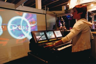 Das Bild zeigt eine junge Frau, die konzentriert an einem professionellen Medienpult arbeitet. Vor ihr befindet sich ein modernes Mischpult mit mehreren integrierten Bildschirmen, die visuelle Darstellungen und Steuerungsoptionen zeigen. Im Hintergrund ist eine große Projektionsfläche zu sehen, auf der bunte, dynamische Grafiken projiziert werden, was auf eine kreative Licht- und Videoproduktion hinweist. Die Umgebung deutet auf eine Messe oder Ausstellung hin, mit technischer Ausstattung und einem Fokus auf Licht- und Medientechnik. Die Frau trägt einen hellen Pullover und scheint mitten in einer technischen Demonstration oder Performance zu sein.