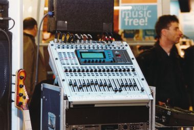Das Bild zeigt ein professionelles Audiomischpult, das auf einem stabilen Rack montiert ist. Die Konsole verfügt über zahlreiche Regler, Schieberegler und Anschlüsse, die eine präzise Steuerung des Tons ermöglichen. Oben auf dem Mischpult sind Kabel eingesteckt, die den Eindruck einer aktiven Nutzung vermitteln. Im Hintergrund ist eine Umgebung mit Personen zu erkennen. Links unten ist der Kopf eines Basses zu sehen, der auf die musikalische Ausrichtung der Präsentation hinweist. Ein Schild im Hintergrund trägt den Text „your music free!“, was auf eine werbliche oder promotive Umgebung schließen lässt.