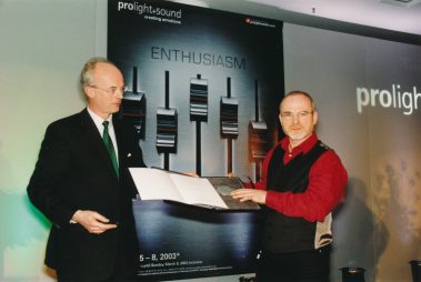 Das Bild wurde im Rahmen der Prolight + Sound 2003 aufgenommen. Im Mittelpunkt stehen zwei Männer: Der links abgebildete Mann trägt einen dunklen Anzug mit grünem Schlips und hält eine Mappe, während der rechts abgebildete Mann mit Brille, rotem Hemd und schwarzer Weste eine Urkunde oder einen Preis entgegennimmt. Im Hintergrund ist ein großes Plakat mit dem Schriftzug „Prolight + Sound“ und dem Slogan „creating emotions“ zu sehen, ergänzt durch das Wort „ENTHUSIASM“. Das stilisierte Design mit Regler-Motiven unterstreicht den Fokus der Messe auf Licht- und Tontechnik. Die Szene vermittelt eine professionelle und feierliche Atmosphäre.