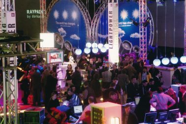 Das Bild zeigt eine lebendige Messeszene mit vielen Besuchern, die sich an verschiedenen Ständen und Displays versammeln. Große Werbebanner mit Aufschriften wie „Lightpower“ und „The world has changed“ heben das Thema Licht- und Veranstaltungstechnik hervor. Im Hintergrund sind auffällige Dekorationen wie beleuchtete Kugeln und große Lautsprecher-Attrappen zu sehen, während vorne mehrere Monitore und technische Geräte die Aufmerksamkeit auf sich ziehen. Die bunte Beleuchtung und die dichten Menschenmengen vermitteln eine dynamische und innovative Atmosphäre, typisch für eine Fachmesse dieser Art.