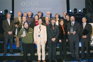 Das Bild zeigt eine Gruppe von Preisträgern und Beteiligten bei der Verleihung des „Opus – Deutscher Bühnenpreis“. Die Personen posieren gemeinsam auf einer Bühne vor einer Leinwand mit dem Schriftzug des Preises. Einige halten Auszeichnungen in der Hand, was auf ihre Leistungen im Bereich Bühnen- und Veranstaltungstechnik hinweist. Die Anwesenden sind unterschiedlich gekleidet – von formellen Anzügen bis zu legereren Outfits, was die Vielfalt der Teilnehmer unterstreicht. Die Atmosphäre wirkt feierlich und professionell, mit einer klaren Ausleuchtung und Bühnenelementen im Hintergrund, die den Rahmen der Preisverleihung abrunden.