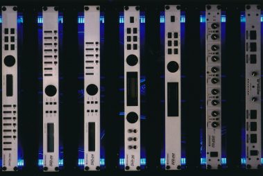 Das Bild zeigt eine Reihe von professionellen Audio-Rackgeräten, die in einem Regal mit blauer Hintergrundbeleuchtung montiert sind. Die Gerätefrontplatten sind in einem einheitlichen, minimalistischen Design gehalten, mit Bedienelementen wie Drehreglern, Tasten und LED-Anzeigen. Jedes Gerät trägt unterschiedliche Beschriftungen und Modellnamen, was auf eine Vielzahl von Funktionen und Anwendungen hinweist. Die symmetrische Anordnung und die Beleuchtung verleihen der Präsentation einen modernen und technologischen Charakter. 