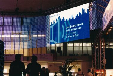 Das Bild zeigt eine großformatige LED-Wand auf der Prolight + Sound, die Informationen über das „Live Sound Forum“ anzeigt. Die Veranstaltung findet in der Halle 9 im Außenbereich statt, wie auf der blauen Anzeige mit weißer Schrift vermerkt ist, die auch Datum und Uhrzeiten angibt. Die LED-Wand ist Teil einer beeindruckenden Lichtinstallation mit gebogenen LED-Panels, die in verschiedenen Farben leuchten und die Szenerie modern und technologisch wirken lassen. Im Vordergrund sind die Silhouetten von Messebesuchern zu sehen, die sich vor der Wand bewegen. Die Szenerie vermittelt eine lebhafte und innovative Atmosphäre, typisch für eine Messe mit Fokus auf Veranstaltungstechnik.