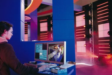 Das Bild zeigt eine Person, die an einem Computerarbeitsplatz auf einer Messe arbeitet. Vor ihr stehen zwei Monitore, die ein Lichtdesign-Programm darstellen, mit dem offenbar Bühnenelemente und Beleuchtung simuliert werden. Im Hintergrund sind eine aufwändig beleuchtete Wand mit LED-Streifen und Scheinwerfern sowie weitere Messestand-Elemente zu sehen. Die Szenerie ist in kräftigen Farben gehalten, vor allem Blau und Rot, was die technische und dynamische Atmosphäre unterstreicht. Der Arbeitsplatz vermittelt den Eindruck eines Live-Demonstrationsbereichs, der den Einsatz von Software für Bühnen- und Veranstaltungstechnik präsentiert.