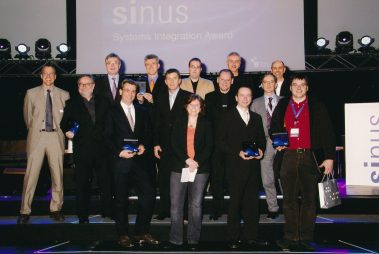Das Bild zeigt die Gewinner der Verleihung des „Sinus – Systems Integration Award“. Die Gruppe, bestehend aus Männern und einer Frau, posiert auf einer Bühne vor einer großen Leinwand mit dem Schriftzug des Preises. Einige halten die Auszeichnungen in Form von eleganten Trophäen in ihren Händen. Die Kleidung variiert von formellen Anzügen bis zu etwas legereren Outfits, was die Vielfalt der Beteiligten unterstreicht. Im Hintergrund sind Bühnenbeleuchtung und technische Elemente zu sehen, die die feierliche und professionelle Atmosphäre der Veranstaltung hervorheben. Die Stimmung wirkt stolz und wertschätzend.