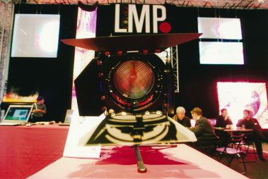 Das Bild zeigt einen Messestand des Unternehmens „LMP“ mit einem großen Scheinwerfer im Vordergrund, der als zentraler Blickfang präsentiert wird. Der Scheinwerfer ist frontal ausgerichtet, wodurch die detaillierte Fresnellinse und die Blendenflügel gut sichtbar sind. Im Hintergrund ist das LMP-Logo auf einem schwarzen Banner zu erkennen, flankiert von bunten LED-Displays, die für visuelle Dynamik sorgen. Weiter hinten sind Messebesucher in Gespräche vertieft, die an Tischen sitzen. Die Szene vermittelt eine professionelle und innovative Atmosphäre, die den Fokus auf Beleuchtungstechnologie und deren praktische Anwendung legt.