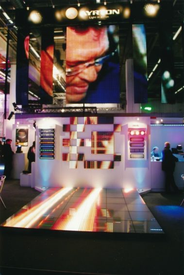 Das Bild zeigt einen eindrucksvollen Messestand des Unternehmens „Ayrton“, spezialisiert auf Beleuchtungstechnologie. Im Mittelpunkt steht ein beleuchteter Boden mit LED-Effekten, der durch fließende Lichtstrahlen dynamisch wirkt. Dahinter sind eine Wand mit modularen LED-Panels und darüber mehrere vertikale LED-Displays zu sehen, die ein übergroßes Porträt zeigen. Der gesamte Stand ist mit intensiven Lichteffekten gestaltet und von modernem Design geprägt. Der Schriftzug „Ayrton – Light in Motion“ über dem Stand hebt die Marke hervor. Im Hintergrund sind Messebesucher und weitere Lichtinstallationen erkennbar, die eine lebendige und technische Atmosphäre schaffen.