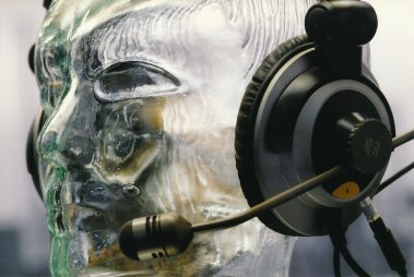 Das Bild zeigt eine Nahaufnahme eines transparenten, gläsernen Kopfmodells, das ein Headset trägt. Das Headset besteht aus großen Ohrmuscheln und einem Mikrofonarm, der sich vor dem Mundbereich des Modells befindet. Die Details des Kopfhörers und des Mikrofons sind präzise sichtbar, einschließlich Kabelverbindungen und Einstellreglern. Die Wahl des gläsernen Kopfes hebt das Design und die technische Präzision des Headsets hervor. Die Aufnahme wirkt professionell, um die Funktionalität und Ästhetik des Produkts zu demonstrieren.