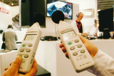 Das Bild zeigt zwei Schallpegelmessgeräte, die von unterschiedlichen Personen gehalten werden. Die Geräte messen den aktuellen Schalldruckpegel, der auf den Displays zu sehen ist (82,0 und 82,1 dB). Im Hintergrund befinden sich ein DJ mit Kopfhörern und eine große Lautsprecherbox, was darauf hindeutet, dass die Messung in einer Messeumgebung mit musikalischer Beschallung stattfindet. Ein Monitor zeigt ein Nahbild technischer Geräte, was den Fokus auf Audiotechnik und Präzision unterstreicht. Die Szene vermittelt eine professionelle und technische Atmosphäre, bei der Schallmessung als Teil der Veranstaltungstechnik präsentiert wird.