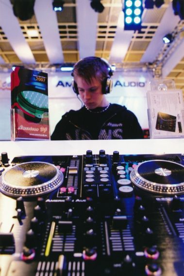 Das Bild zeigt einen jungen DJ, der an einem professionellen DJ-Controller arbeitet. Er trägt Kopfhörer und konzentriert sich auf seine Tätigkeit, während er vor einem beleuchteten Messestand steht. Im Vordergrund ist das Mischpult mit Reglern, Knöpfen und Jog-Wheels gut sichtbar. Auf dem Tisch befinden sich außerdem Werbematerialien, darunter ein Flyer mit der Aufschrift „American DJ 2007“, was auf den Hersteller und das Jahr der Präsentation hinweist. Im Hintergrund sind Beleuchtungselemente und ein Banner mit dem Schriftzug „American Audio“ zu erkennen, die die Szene als eine Vorführung auf einer Messe oder Veranstaltung für Audiotechnik kennzeichnen. Die Atmosphäre wirkt modern und technologieorientiert.