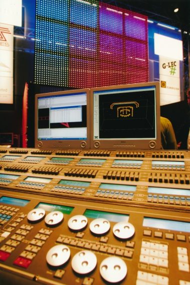 Das Bild zeigt ein modernes Lichtsteuerpult mit mehreren Reihen von Reglern, Tasten und Displays, die für die Steuerung komplexer Beleuchtungssysteme verwendet werden. Im Hintergrund ist eine große LED-Wand zu sehen, die in bunten Farben leuchtet und ein Raster aus Lichtpunkten darstellt. Zwei Monitore auf dem Pult zeigen grafische Benutzeroberflächen, die vermutlich die Steuerung und Visualisierung der Lichtinstallation ermöglichen. Umgeben von Logos und Bannern wirkt die Szene wie ein technischer Messestand, der innovative Lösungen im Bereich der Lichttechnik präsentiert. Die Kombination aus moderner Technologie und visueller Darstellung vermittelt einen professionellen und futuristischen Eindruck.