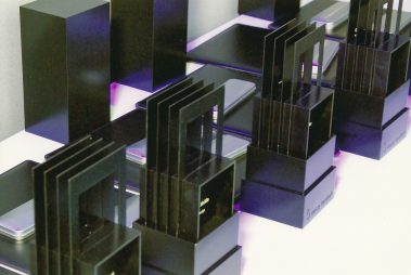 Das Bild zeigt eine Reihe von modernen Trophäen, die für eine Preisverleihung vorbereitet wurden. Die Awards bestehen aus schwarzen geometrischen Elementen, die eine klare, minimalistische Formensprache aufweisen. Auf den Sockeln ist das Logo der Messe Frankfurt zu erkennen. Die Trophäen sind symmetrisch aufgestellt, und die Szene ist in lila-weißem Licht beleuchtet, wodurch die Objekte elegant und hochwertig wirken.