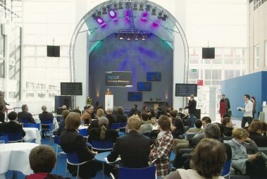 Das Bild zeigt eine Preisverleihung oder Präsentation im Rahmen des „Opus – Deutscher Bühnenpreis“. Im Mittelpunkt steht eine Bühne mit einem Rednerpult, hinter dem eine Person spricht. Die Bühne ist mit einer Beleuchtung in Blau- und Lilatönen ausgeleuchtet, flankiert von Bildschirmen, die ergänzende Informationen anzeigen. Vor der Bühne sitzen zahlreiche Zuschauer an Tischen mit weißen Tischdecken und blauen Stühlen. Der Veranstaltungsraum wirkt offen und lichtdurchflutet, mit großen Glasfronten im Hintergrund. Es herrscht eine professionelle, aber gleichzeitig entspannte Atmosphäre.