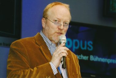 Das Bild zeigt einen Mann mittleren Alters, der in ein Mikrofon spricht, vermutlich während einer Präsentation oder Moderation. Er trägt ein braunes Cord-Sakko, ein kariertes Hemd und eine Brille. Im Hintergrund ist unscharf ein Bildschirm zu erkennen, auf dem das Wort „Opus“ und „Deutscher Bühnenpreis“ zu lesen sind. Dies deutet darauf hin, dass die Szene im Rahmen einer Veranstaltung oder Preisverleihung mit Bezug zur Bühnentechnik stattfindet. Der Mann wirkt konzentriert und in die Moderation vertieft.