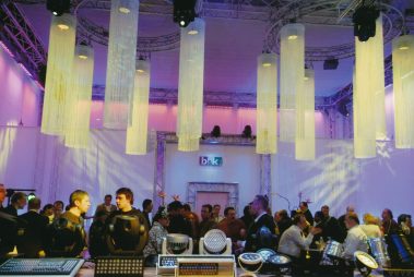 Das Bild zeigt eine lebhafte Messe- oder Veranstaltungsszene, in der eine größere Gruppe von Menschen in einer modern gestalteten Halle zusammenkommt. Im Vordergrund sind verschiedene Beleuchtungsgeräte und ein Mischpult zu sehen, die Teil der Ausstellung für Veranstaltungstechnik sind. Über der Menge hängen große, leuchtende Lampenschirme in zylinderförmigem Design, die eine stimmungsvolle Atmosphäre schaffen. Der Raum ist mit Traversen, buntem Licht in Rosa- und Blautönen sowie technischen Elementen ausgestattet. Im Hintergrund ist ein Schild mit der Aufschrift „b&k“ sichtbar. Die Anwesenden wirken beschäftigt, viele sind in Gespräche vertieft.