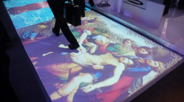 Das Bild zeigt einen beleuchteten Boden, auf dem eine Projektion eines klassischen Gemäldes dargestellt wird. Die Projektion scheint religiöse oder historische Motive zu enthalten, mit Figuren in dramatischer Pose und lebhaften Farben. Mehrere Personen laufen über die Projektion, was darauf hindeutet, dass es sich um eine interaktive oder ästhetische Installation auf einer Messe handelt. Die Schuhe der Personen und eine schwarze Aktentasche sind im Bild sichtbar, was den Kontrast zwischen Kunst und Moderne verstärkt. Die Umgebung wirkt futuristisch und innovativ, passend für eine Veranstaltung, die Technologie und Kunst verbindet.