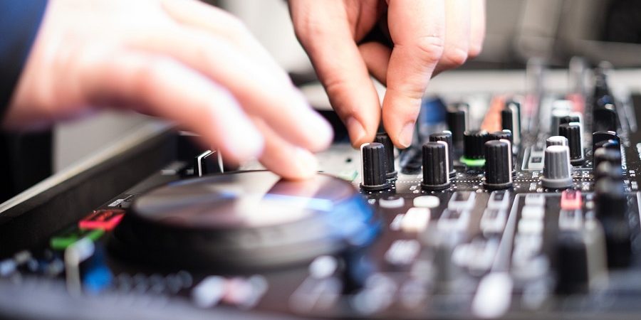 Das Bild zeigt eine Nahaufnahme eines Mischpults, auf dem sich mehrere Drehregler, Schieberegler und Knöpfe befinden. Zwei Hände justieren verschiedene Einstellungen – eine Hand dreht einen Regler, während die andere anscheinend einen anderen Bereich des Mischpults bedient. Die Aufnahme ist leicht unscharf im Vordergrund, wodurch der Fokus auf die Handbewegungen und die Bedienelemente gelegt wird. Das Bild vermittelt eine dynamische Atmosphäre und suggeriert eine intensive Arbeit mit Audiotechnik, wie sie beispielsweise im MixLab auf der Prolight + Sound zum Einsatz kommt.