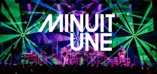Das Bild zeigt eine beeindruckende Live-Bühnenszene mit einer dynamischen Lichtshow. Der Hintergrund ist von intensiven grünen und violetten Lichteffekten geprägt, die strahlenförmig von verschiedenen Punkten ausgehen und den gesamten Raum einhüllen. In der Mitte des Bildes befindet sich das große, weiße Logo von Minuit Une, das prominent über die Szene gelegt wurde. Auf der Bühne stehen mehrere Musiker, die von einer aufwendigen Lichtinstallation umgeben sind. Die Beleuchtung erzeugt eine immersive Atmosphäre, die typisch für die IVL-Technologie von Minuit Une ist. Die Kombination aus multidirektionalen Lichtstrahlen, Projektionen und der energiegeladenen Live-Performance vermittelt den Eindruck eines intensiven, mitreißenden Konzerterlebnisses.