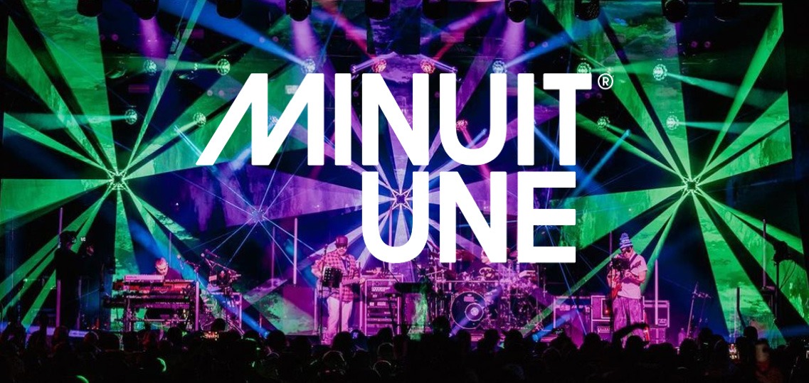 Das Bild zeigt eine beeindruckende Live-Bühnenszene mit einer dynamischen Lichtshow. Der Hintergrund ist von intensiven grünen und violetten Lichteffekten geprägt, die strahlenförmig von verschiedenen Punkten ausgehen und den gesamten Raum einhüllen. In der Mitte des Bildes befindet sich das große, weiße Logo von Minuit Une, das prominent über die Szene gelegt wurde. Auf der Bühne stehen mehrere Musiker, die von einer aufwendigen Lichtinstallation umgeben sind. Die Beleuchtung erzeugt eine immersive Atmosphäre, die typisch für die IVL-Technologie von Minuit Une ist. Die Kombination aus multidirektionalen Lichtstrahlen, Projektionen und der energiegeladenen Live-Performance vermittelt den Eindruck eines intensiven, mitreißenden Konzerterlebnisses.