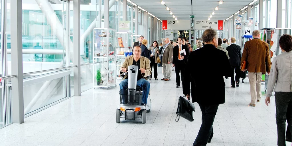 Ein Mann fährt mit einem Elektroscooter durch einen barrierefreien Messegang mit vielen Besuchenden.
