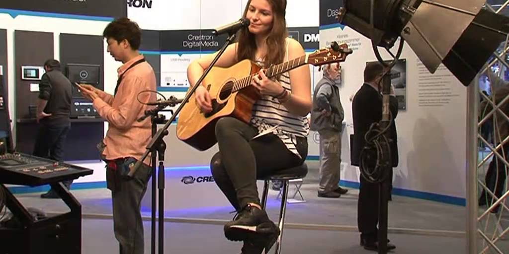 Bewegte Impressionen von der Prolight + Sound 2015 Das Bild zeigt eine junge Frau, die auf einem Barhocker sitzt und eine Akustikgitarre spielt. Sie lächelt leicht, während sie ins Mikrofon singt, das vor ihr positioniert ist. Um sie herum ist eine Messeumgebung zu sehen, mit Informationsständen und Besuchern im Hintergrund. Eine professionelle Beleuchtung, bestehend aus einem großen Scheinwerfer und weiteren technischen Geräten, hebt die Performerin hervor. Links im Bild steht ein Mann, der auf sein Handy schaut, während im Hintergrund weitere Messebesucher die ausgestellten Produkte und Displays betrachten. Die Szene vermittelt eine Mischung aus musikalischer Darbietung und technischer Präsentation, typisch für eine Veranstaltung wie die Prolight + Sound.