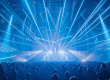 A large crowd stands in a concert hall and looks up at a stage above which numerous powerful blue and white beams of light shoot out into the room in a fan shape. The light show creates a dense network of laser and spot beams that fill the entire hall and create a dynamic, energetic atmosphere.