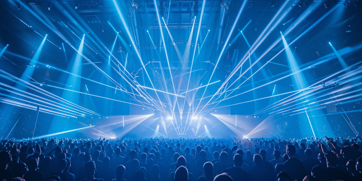 A large crowd stands in a concert hall and looks up at a stage above which numerous powerful blue and white beams of light shoot out into the room in a fan shape. The light show creates a dense network of laser and spot beams that fill the entire hall and create a dynamic, energetic atmosphere. 