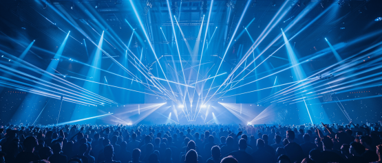A large crowd stands in a concert hall and looks up at a stage above which numerous powerful blue and white beams of light shoot out into the room in a fan shape. The light show creates a dense network of laser and spot beams that fill the entire hall and create a dynamic, energetic atmosphere. 