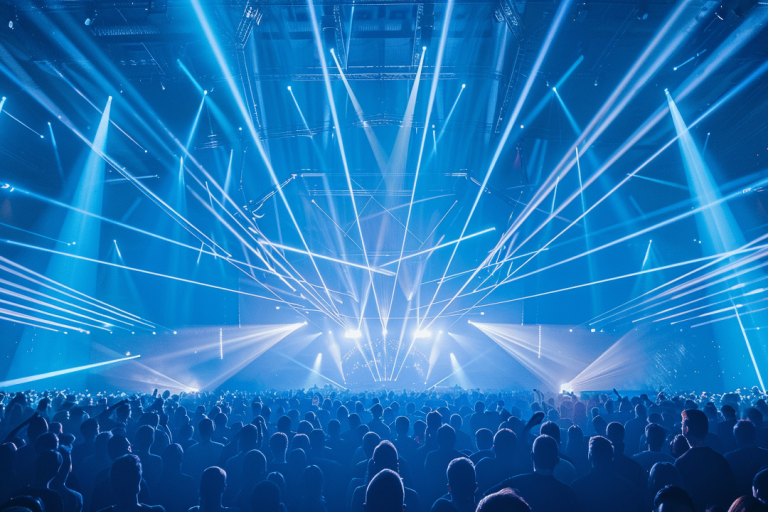 A large crowd stands in a concert hall and looks up at a stage above which numerous powerful blue and white beams of light shoot out into the room in a fan shape. The light show creates a dense network of laser and spot beams that fill the entire hall and create a dynamic, energetic atmosphere. 