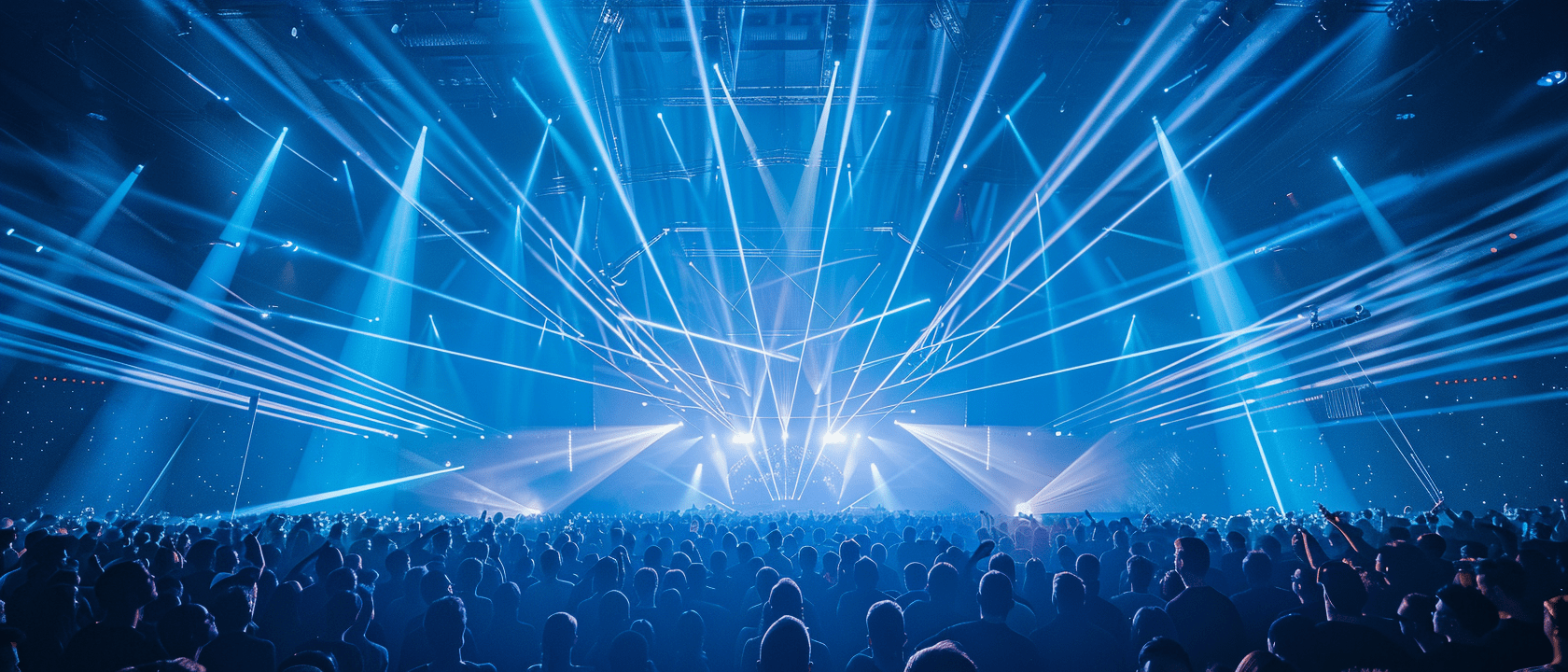 A large crowd stands in a concert hall and looks up at a stage above which numerous powerful blue and white beams of light shoot out into the room in a fan shape. The light show creates a dense network of laser and spot beams that fill the entire hall and create a dynamic, energetic atmosphere. 