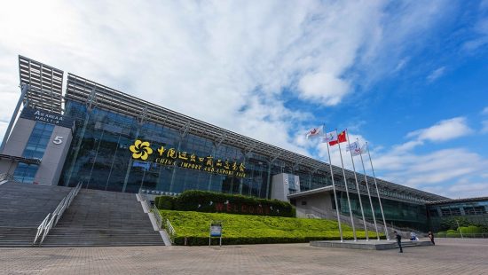 Modern glass façade of the China Import & Export Fair Complex with stairs, flagpoles and blue sky.