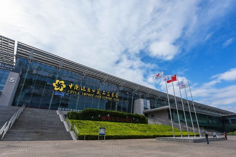 Modern glass façade of the China Import & Export Fair Complex with stairs, flagpoles and blue sky.