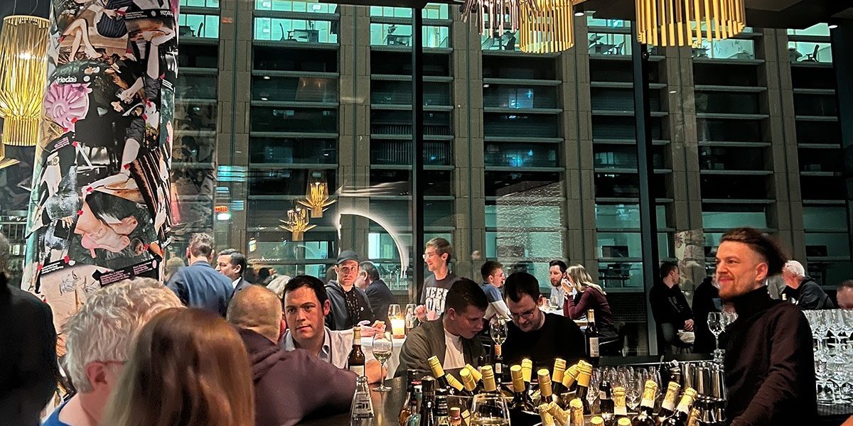 community-nights-pls-2024 Das Bild zeigt eine lebhafte und moderne Barszene, vermutlich von einem Networking-Event oder einer Community-Veranstaltung, bei der Menschen in entspannter Atmosphäre miteinander ins Gespräch kommen. Im Mittelpunkt steht ein großer Tresen, reich dekoriert mit Flaschen und Gläsern, während Teilnehmer in Gespräche vertieft sind oder das Ambiente genießen. Auffällige Designelemente, wie eine mit Collagen gestaltete Säule und stilvolle Beleuchtung, prägen den Raum, der durch große Fensterfronten den Blick auf ein modernes Gebäude freigibt. Die warmen Lichtakzente und die gelöste Stimmung vermitteln den Eindruck eines gelungenen Abends, der Begegnung und Austausch fördert.