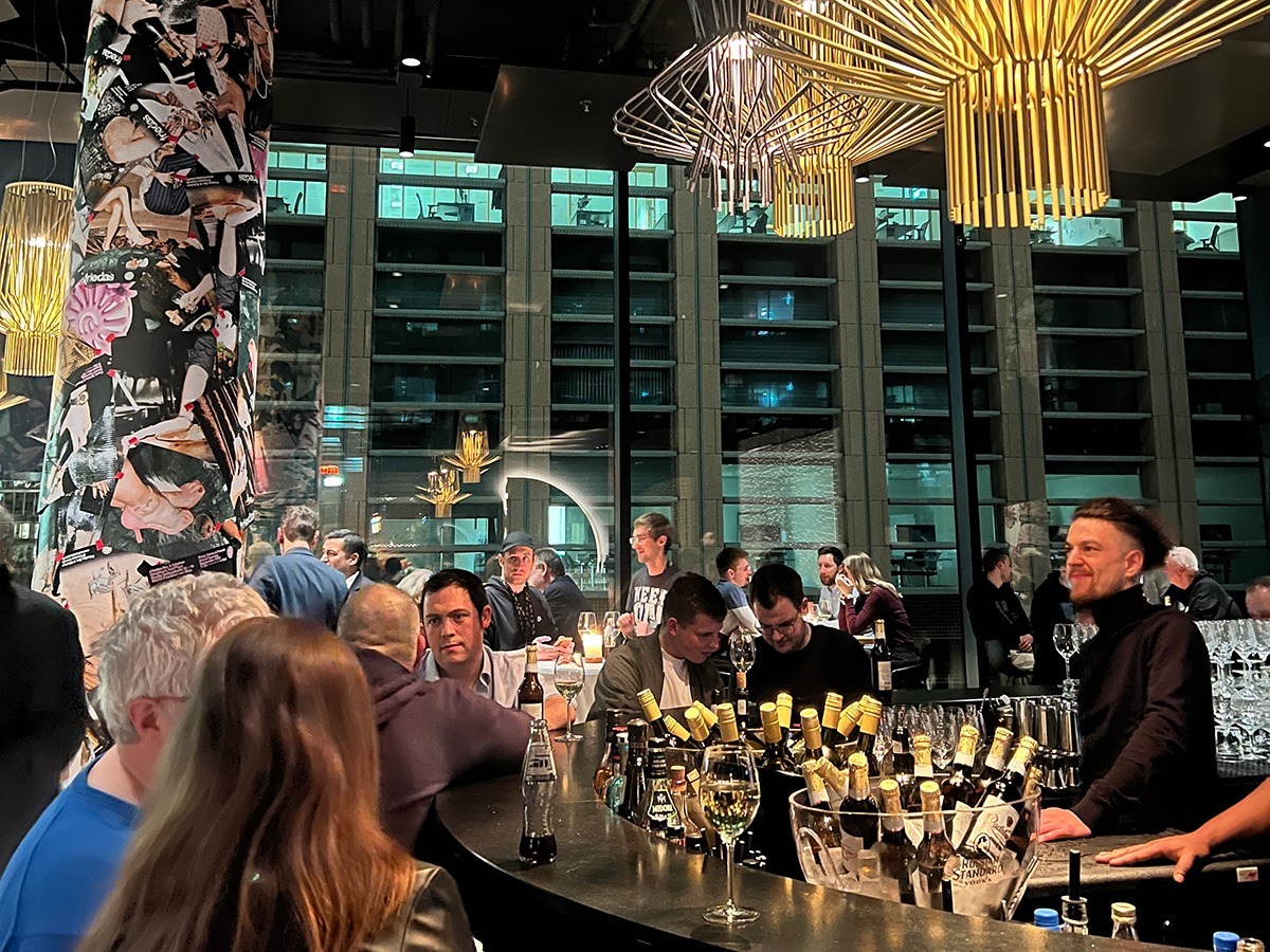 Das Bild zeigt eine lebhafte und moderne Barszene, vermutlich von einem Networking-Event oder einer Community-Veranstaltung, bei der Menschen in entspannter Atmosphäre miteinander ins Gespräch kommen. Im Mittelpunkt steht ein großer Tresen, reich dekoriert mit Flaschen und Gläsern, während Teilnehmer in Gespräche vertieft sind oder das Ambiente genießen. Auffällige Designelemente, wie eine mit Collagen gestaltete Säule und stilvolle Beleuchtung, prägen den Raum, der durch große Fensterfronten den Blick auf ein modernes Gebäude freigibt. Die warmen Lichtakzente und die gelöste Stimmung vermitteln den Eindruck eines gelungenen Abends, der Begegnung und Austausch fördert.