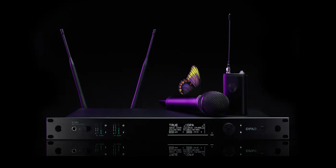 dpa-n-series-wireless Das Bild zeigt ein hochwertiges Drahtlossystem aus der DPA N-Serie Digital Wireless, bestehend aus mehreren Komponenten. Im Mittelpunkt steht ein N-DR1 Digital Wireless Receiver, ein professioneller Zweikanalempfänger mit zwei aufrecht stehenden Antennen, mehreren LED-Anzeigen und einem Display, das technische Informationen wie Frequenzen und Signalstärke darstellt.
Auf dem Empfänger liegt ein kabelloses Handmikrofon, das zum DPA d:facto™-Sortiment gehören könnte. Rechts daneben steht ein Bodypack-Transmitter mit einer angeschlossenen Antenne, der eine kompakte und robuste Bauweise aufweist. Das gesamte Equipment ist in einem eleganten, schwarzen Design mit modernen, lila Akzenten gehalten.
Besonders auffällig ist ein Schmetterling, der auf dem Mikrofon platziert ist. Dies könnte symbolisch für die klare und natürliche Klangqualität der DPA-Produkte stehen. Der Hintergrund ist tiefschwarz, was das Equipment in den Fokus rückt und die hochwertige Ästhetik der Produktreihe unterstreicht.