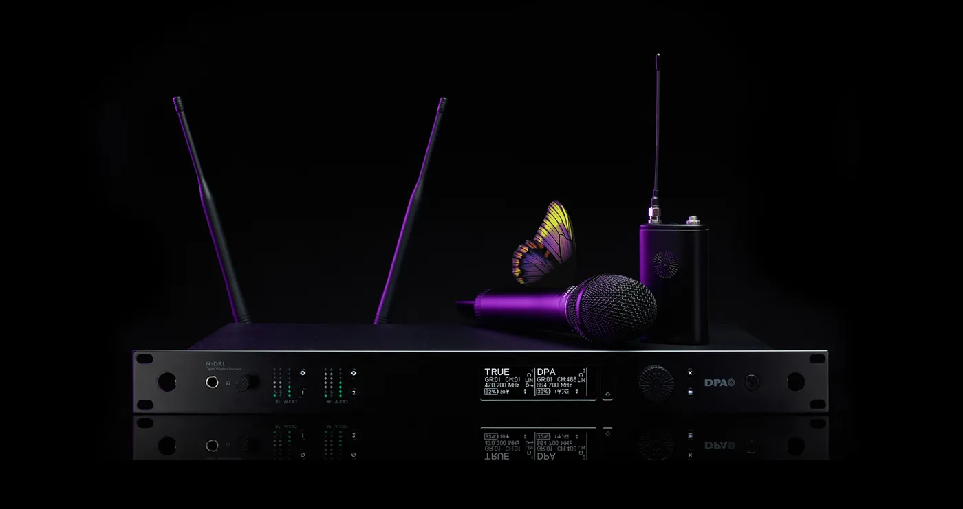 Das Bild zeigt ein hochwertiges Drahtlossystem aus der DPA N-Serie Digital Wireless, bestehend aus mehreren Komponenten. Im Mittelpunkt steht ein N-DR1 Digital Wireless Receiver, ein professioneller Zweikanalempfänger mit zwei aufrecht stehenden Antennen, mehreren LED-Anzeigen und einem Display, das technische Informationen wie Frequenzen und Signalstärke darstellt.

Auf dem Empfänger liegt ein kabelloses Handmikrofon, das zum DPA d:facto™-Sortiment gehören könnte. Rechts daneben steht ein Bodypack-Transmitter mit einer angeschlossenen Antenne, der eine kompakte und robuste Bauweise aufweist. Das gesamte Equipment ist in einem eleganten, schwarzen Design mit modernen, lila Akzenten gehalten.

Besonders auffällig ist ein Schmetterling, der auf dem Mikrofon platziert ist. Dies könnte symbolisch für die klare und natürliche Klangqualität der DPA-Produkte stehen. Der Hintergrund ist tiefschwarz, was das Equipment in den Fokus rückt und die hochwertige Ästhetik der Produktreihe unterstreicht.