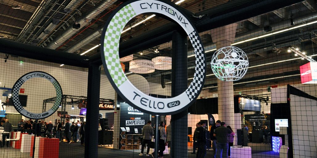 dronen-soccer-pls-2025 Das Bild zeigt ein Drone-Soccer-Spielfeld auf einer Messe oder Eventfläche. Im Vordergrund ist ein großer Torbogen mit der Aufschrift „CYTRONIX TELLO EDU“ zu sehen, gestaltet im typischen Schachbrettmuster. Dahinter fliegt eine Drohne in einem weißen Schutzkäfig – scheinbar auf dem Weg zum Tor oder in einer Spielszene. Um das Spielfeld herum ist ein feinmaschiges Sicherheitsnetz gespannt, das Zuschauerinnen und Zuschauer schützt. Im Hintergrund erkennt man Messestände, leuchtende Displays und Besuchergruppen, was auf eine dynamische, technikorientierte Veranstaltung hinweist. Die Szene verbindet technische Präzision mit interaktivem Messeerlebnis.