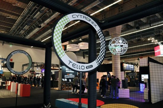 Das Bild zeigt ein Drone-Soccer-Spielfeld auf einer Messe oder Eventfläche. Im Vordergrund ist ein großer Torbogen mit der Aufschrift „CYTRONIX TELLO EDU“ zu sehen, gestaltet im typischen Schachbrettmuster. Dahinter fliegt eine Drohne in einem weißen Schutzkäfig – scheinbar auf dem Weg zum Tor oder in einer Spielszene. Um das Spielfeld herum ist ein feinmaschiges Sicherheitsnetz gespannt, das Zuschauerinnen und Zuschauer schützt. Im Hintergrund erkennt man Messestände, leuchtende Displays und Besuchergruppen, was auf eine dynamische, technikorientierte Veranstaltung hinweist. Die Szene verbindet technische Präzision mit interaktivem Messeerlebnis.