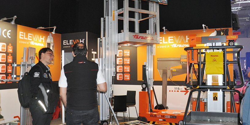 faraone-pls-2025 Das Bild zeigt den Messestand von Faraone auf der Prolight + Sound 2025. Im Zentrum stehen drei verschiedene Modelle der Elevah-Serie: gut sichtbar ist vorne rechts die Elevah 80 Move, rechts dahinter die Elevah 80 ES Move mit ausfahrbarem Arbeitskorb, und im linken Bildbereich die Elevah 70, ein kompakter Personenlift mit manueller Verschiebung. Zwei Besucher unterhalten sich direkt vor dem Elevah 70, während im Hintergrund ein Bildschirm eine Produktanimation zeigt. Der Stand ist in den typischen Farben Grau, Orange und Weiß gestaltet und vermittelt mit technischen Daten an den Rückwänden einen informativen Überblick über die Faraone-Produkte.