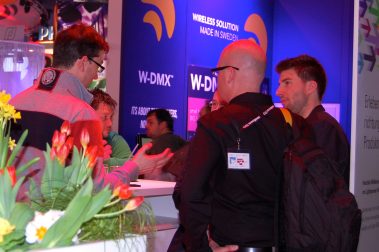 Das Bild zeigt eine kleine Gruppe von Personen, die in einer lebhaften Messeumgebung in ein Gespräch vertieft sind. Im Vordergrund sind Tulpen und Narzissen zu sehen, die eine freundliche und einladende Atmosphäre schaffen. Die Teilnehmer tragen Namensschilder und scheinen in einem fachlichen Austausch zu stehen. Im Hintergrund befinden sich Werbetafeln mit den Logos von "W-DMX" und anderen Marken, die auf die Themen Beleuchtung und Technologie hinweisen. Die Beleuchtung des Standes ist in Blau- und Rottönen gehalten, was der Szene ein modernes und dynamisches Ambiente verleiht. Die Szene vermittelt ein Bild von professionellem Networking und Dialog in einem Messekontext.