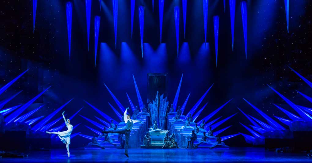 Several dancers perform on a stage bathed in intense blue light, in front of a large, ice-like crystal backdrop. Long, glowing icicles hang above them, protruding into the room. Numerous pointed, glittering elements decorate the stage area, creating a wintry, frosty landscape. The dancers move elegantly through this fairytale ice world.<br>