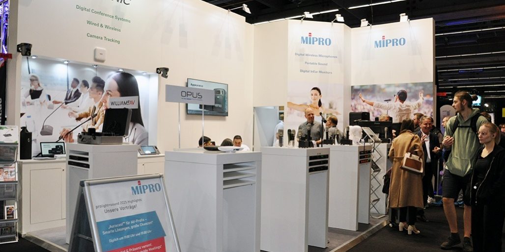 Der Messestand von MIPRO auf der Prolight + Sound 2025 präsentiert sich hell, offen und professionell. Im Zentrum stehen mehrere Produktinseln mit digitaler Drahtlostechnik – darunter Mikrofone, Taschensender und In-Ear-Systeme. Ein aufgestelltes Rollup kündigt spannende Vorträge zu digitalen Mikrofonsystemen, smarter Bühnenpraxis und erfolgreicher Medientechnik an. Die große Besucherzahl am Stand sowie das rege Interesse an den präsentierten Lösungen unterstreichen die Relevanz von MIPRO im professionellen Audiomarkt. Begleitet wird die Ausstellung von auffälligen Visuals, die die Einsatzbereiche der Technik – von Business bis Bühne – eindrucksvoll in Szene setzen.